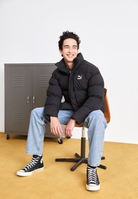 Doudoune noire avec col haut et logo, associée à un jean bleu clair portant un texte blanc, et des baskets montantes noires sur un sol jaune.