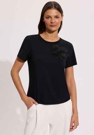 Kobieta ubrana w czarną bluzkę z krótkim rękawem, ozdobioną teksturowanym czarnym kwiatem na piersi, oraz białe spodnie, stoi z jedną ręką w kieszeni.