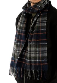 Muž v černé zipové bundě a modro-šedo-oranžové kostkované šále se třásněmi omotaným kolem krku.