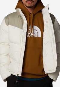 Uppblåsbar jacka i vitt med ljusgröna detaljer, med dragkedjestängning och hög krage. Lager på en brun hoodie med logotyp.