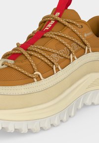 Gros plan d'une chaussure d'extérieur marron et beige avec des semelles blanches épaisses, des lacets à motifs et une languette rouge vif sur la languette.