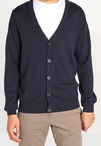 Laivastonsininen neuletakki, jossa on V-pääntie, napitettava etuosa, ribatoidut hihansuut ja helma sekä sileä pinta, kerroksittain valkoisen t-paidan päällä.