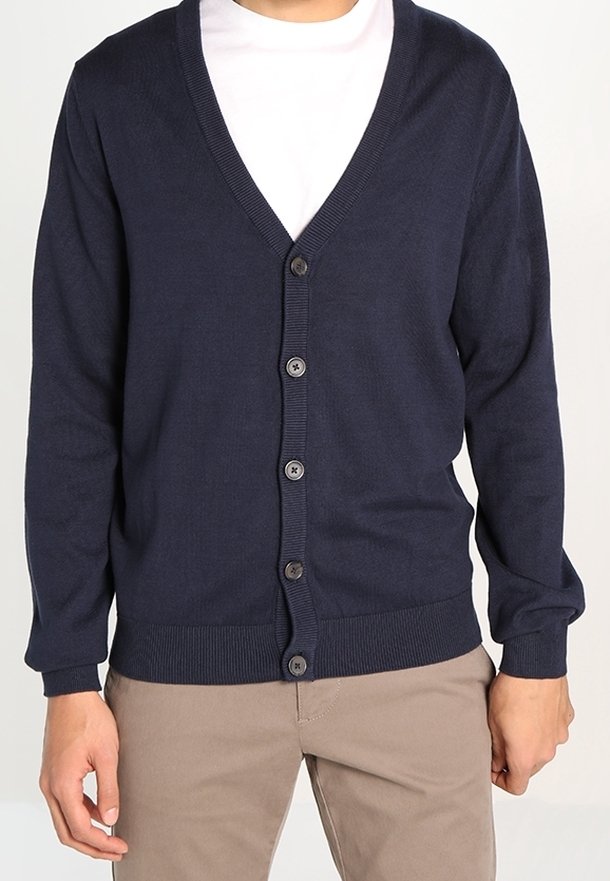 Laivastonsininen neuletakki, jossa on V-pääntie, napitettava etuosa, ribatoidut hihansuut ja helma sekä sileä pinta, kerroksittain valkoisen t-paidan päällä.