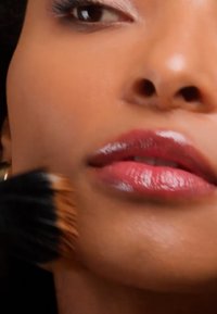 Gros plan sur la partie inférieure du visage d'une personne aux lèvres brillantes appliquant un fond de teint avec un pinceau sur sa joue.