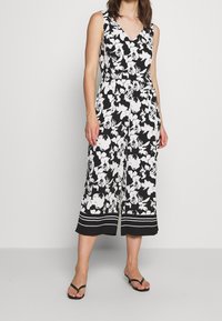 Mujer vistiendo un mono sin mangas de flores en blanco y negro con pantalones de pierna ancha, escote en V y sandalias negras de tacón bajo.