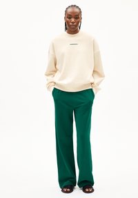 Beige, übergroßer Sweatshirt mit grüner Schrift, kombiniert mit weiten, grünen Hosen. Das Model steht mit Händen in den Taschen und trägt schwarze Sandalen.