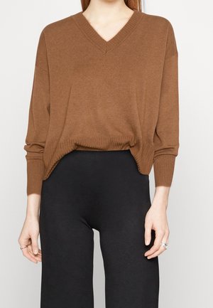Femme portant un pull marron ample à col en V et un pantalon noir, les mains détendues le long du corps, teint clair, cheveux longs roux visibles.