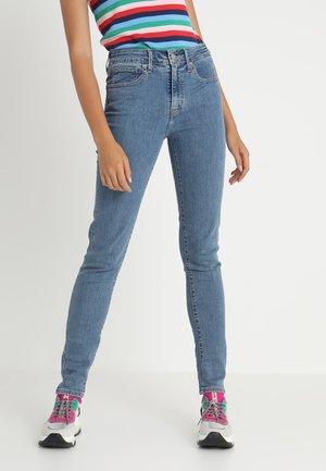 Jeans ajustados de color azul hechos de mezclilla, con cintura alta y cinco bolsillos. Combinados con una blusa a rayas de colores y zapatillas deportivas.