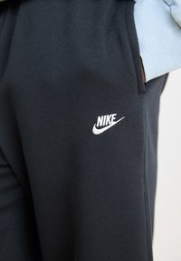Svarta sweatpants i mjukt material med en vit Nike-logotyp på vänster lår och muddar med ljusblå accenter.