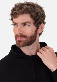 Schwarzer Kapuzenpullover mit Kordelzug, strukturiertem Stoff und gerippten Bündchen. Das Modell hat lockiges braunes Haar und justiert die Kapuze.