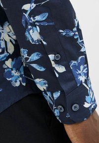 Camicia floreale blu navy con motivi di foglie bianche e azzurre. Polsino con due bottoni abbinati, tessuto liscio con una leggera lucentezza.
