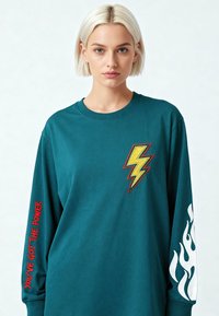 Jeune femme aux cheveux blonds courts portant un t-shirt à manches longues bleu sarcelle avec un éclair jaune, des flammes blanches et le texte rouge "Vous avez le pouvoir" sur les manches.