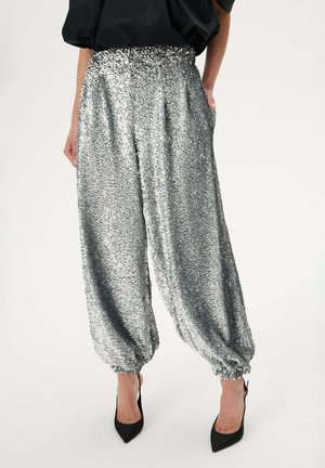 Osoba ubrana w błyszczące srebrne spodnie z cekinami i elastycznymi mankietami, czarną bluzkę oraz czarne szpilki z ostrym noskiem, stojąca na jednolitym tle.