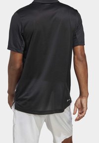 Černé polo tričko s krátkými rukávy a hladkou strukturou, s límečkem a bočními rozparky. Bílé šortky jsou částečně viditelné.