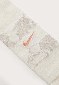 Nike-sokker i en lys cremefarve med et struktureret mønster. Har et orange Nike-logo og subtile grå accenter i hele designet.