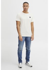 Lysbeige kortærmet t-shirt med et lille sort mærke, parret med blå jeans og grå sneakers. Enkelt design og standard pasform.
