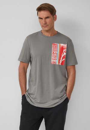 T-shirt gris à manches courtes avec une application graphique colorée comportant du texte et un design abstrait sur le côté gauche. Tissu doux, coupe décontractée.