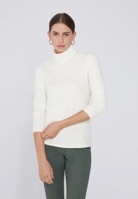 Camisa de cuello alto color beige claro con mangas largas, diseño ajustado y textura suave. Combinada con pantalones verdes, el modelo tiene las manos en las caderas.
