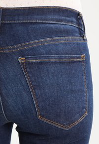 Vaqueros de mezclilla azul oscuro con un diseño ajustado, que presentan costuras naranjas, un bolsillo con parche y una textura suave en la parte posterior.