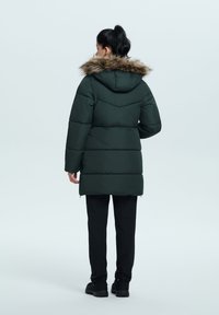 Mørkegrøn pufferjakke med en faux fur hættekant, vandret quilting og sidelommer med lynlås. Båret over sorte bukser og sko.