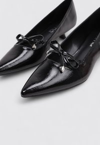 Chaussures en cuir verni noir à bout pointu avec une finition brillante, comportant un nœud décoratif et des accents argentés à l'avant.
