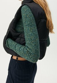 Czarna pikowana kamizelka noszona na zielonym swetrze o fakturze. Kamizelka ma wysoki kołnierz, natomiast sweter długie rękawy i dopasowany krój.