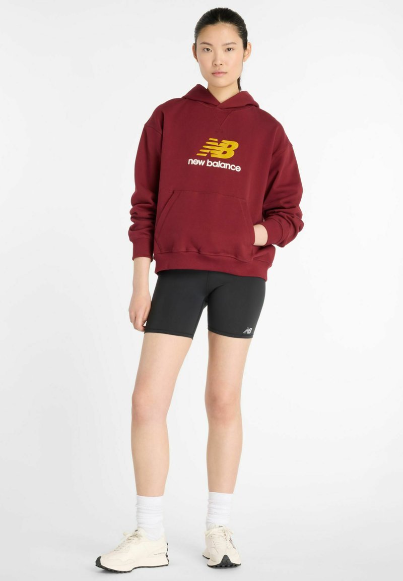 Burgundy hooded sweatshirt with yellow "new balance" logo, kangaroo pocket, paired with black cycling shorts and off-white sneakers.