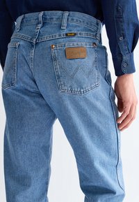 Light blue Wrangler jeans worn by a person with a dark blue shirt, showing back pockets and leather brand patch on the right pocket.