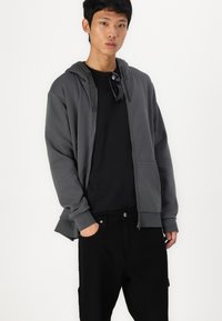 Felpa grigia con zip e coulisse, realizzata in materiale morbido. Indossata sopra una maglietta nera, abbinata a pantaloni neri. Design semplice e vestibilità casual.