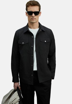 Man draagt een zwart overhemd over een wit T-shirt, zwarte zonnebril, houdt een opgevouwen lichtgekleurde jas in zijn linkerhand.