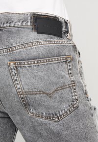 Ljust grå denimjeans med en bakficka som har brun sömnad och en svart läderlogotyp över fickan.