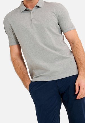 Mand iført en lysegrå kortærmet polo med tre knapper, kombineret med mørkeblå bukser, den ene hånd i lommen.