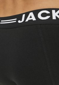 Svart elastisk midjeband med "JACK" i vit stickad text. Tyget är mjukt och elastiskt, och visar en nära bild av plaggets sida.