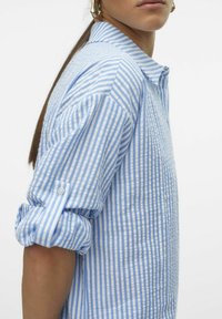 Camicia a righe blu e bianche con colletto, maniche arrotolate e una texture di cotone morbido, che mostra righe verticali e una vestibilità rilassata.