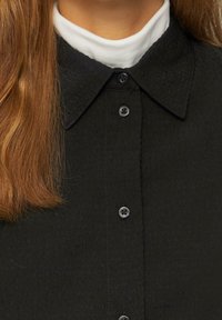Camicia nera con bottone, realizzata in un tessuto testurizzato, con colletto a discesa e quattro bottoni neri sulla parte anteriore. Colletto bianco visibile sotto.