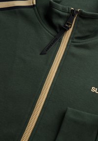 Chaqueta verde con cremallera y cuello alto, que presenta una cremallera crema, un tirador negro y un pequeño logo. Tejido suave con textura mínima.