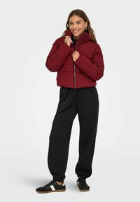 Doudoune bordeaux avec col montant, fermeture éclair et design matelassé, associée à un pantalon de survêtement noir et des baskets noires avec des accents blancs.