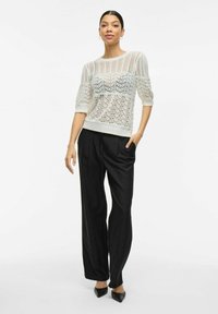 Bílý pletený svetr s krajkovým vzorem, krátké nabírané rukávy, doplněný vysokými černými kalhotami a černými špičatými podpatky.