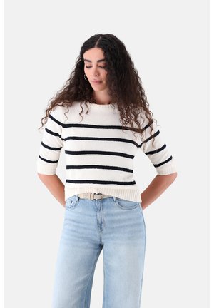 Frau mit lockigem Haar, die einen weißen Pullover mit schwarzen horizontalen Streifen und hellblaue Jeans trägt, die Hände hinter dem Rücken, schaut nach unten.