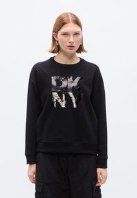 Μαύρο φούτερ από απαλό υλικό, με γυαλιστερό ασημί λογότυπο "DK NY" σε έντονη γραμματοσειρά στο κέντρο. Άνετη εφαρμογή, ριμπ μανσέτες.