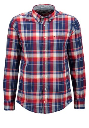 Man wearing a gray crew neck sweater over a red and blue plaid shirt, black slim-fit pants, and black slip-on shoes, standing with one hand in pocket.