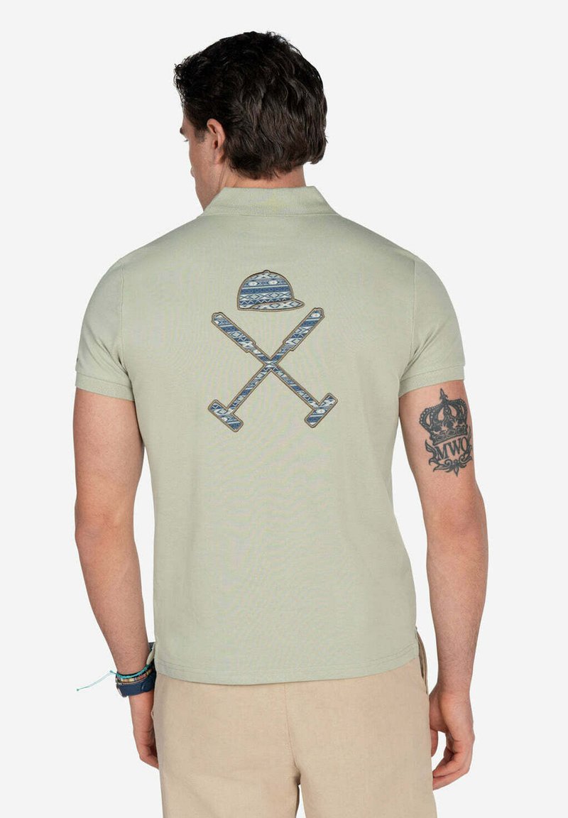 Polo de color verde claro con mangas cortas y un diseño gráfico de un sombrero y palos cruzados en la parte posterior en azul con detalles estampados.
