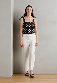 Top fără mâneci alb-negru cu buline, cu bretele legate, purtat cu pantaloni largi albi cu dungi și sandale negre, pe o podea din lemn.