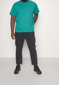 T-shirt verde in cotone con collo rotondo, abbinata a jeans neri a vestibilità dritta e scarpe da ginnastica nere con accenti viola.