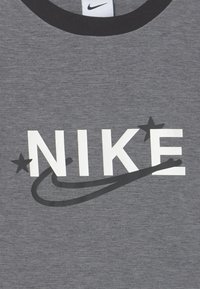 Szara bawełniana koszulka z czarnym okrągłym dekoltem, z dużym białym napisem "NIKE" oraz czarnym graficznym logo w kształcie „swoosh”, z dodatkowymi gwiazdkami podkreślającymi wzór.