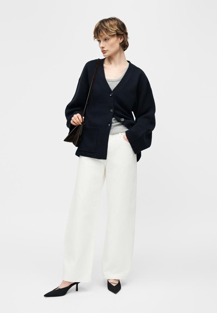 Femme portant un cardigan noir sur un haut gris, un pantalon blanc à jambes larges, des talons noirs, et portant un sac à bandoulière noir, debout devant un fond blanc.