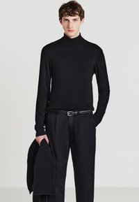 Maglione nero a collo alto in tessuto liscio, abbinato a pantaloni neri e a una sciarpa lavorata a maglia, tenuta in una mano.