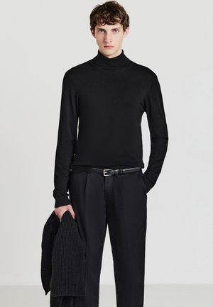 Maglione nero a collo alto in tessuto liscio, abbinato a pantaloni neri e a una sciarpa lavorata a maglia, tenuta in una mano.