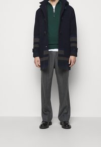 Cappotto di lana blu navy con accenti a strisce, maglione verde con zip, camicia bianca, pantaloni grigi e scarpe nere. Presenta bottoni e tasche.