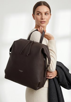 Donna con maglione crema tiene uno zaino grande in pelle marrone scuro sulla spalla e un cappotto grigio scuro piegato in mano.
